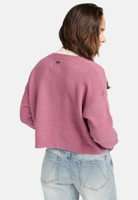 Un pull court rose clair en tissu tricot doux ; présente un ourlet côtelé et un détail d'étiquette au dos. Associe avec un jean bleu clair.