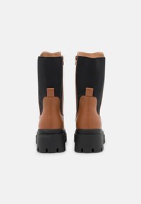 Une paire de bottes mi-mollet avec tige en tissu noir, soutien en cuir brun au niveau du talon, semelles en caoutchouc noir épaisses, et détail de fermeture éclair à l'arrière sur fond blanc.
