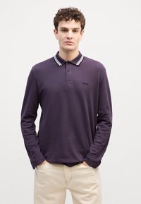 Νεαρός άνδρας με σγουρά μαλλιά, ντυμένος με ένα σκούρο μωβ μακρυμάνικο πόλο μπλουζάκι και ανοιχτό μπεζ παντελόνι, που στέκεται μπροστά σε έναν απλό λευκό τοίχο.