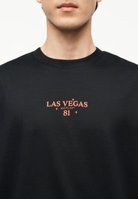 Άνδρας που φοράει μαύρο μπλουζάκι με στρογγυλή λαιμόκοψη, με πορτοκαλί κείμενο "LAS VEGAS REPLAY 81" στο κέντρο του στήθους, σε απλό λευκό φόντο.