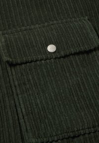 Donkergroen corduroy-stof met een gestructureerd geribbeld patroon, voorzien van een voorzak en een metalen drukknoopsluiting.