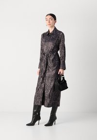Abito camicia con stampa leopardata in tonalità scure, tessuto setoso, colletto, dettaglio cintura legata in vita, maniche lunghe. Modella che porta una borsa nera e indossa stivali con tacco.