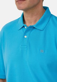 Polo bleu clair avec un col classique, trois boutons et une texture subtile, accompagné d'un petit logo sur la poitrine.