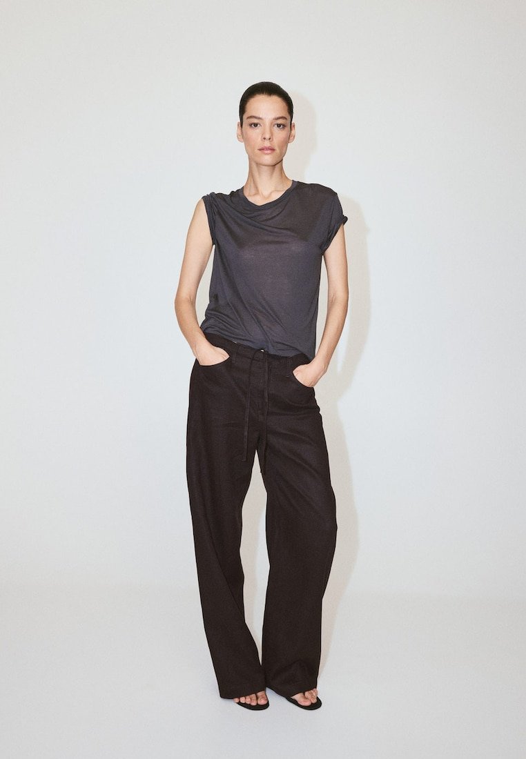 Femme debout sur un fond uni, portant un haut sans manches foncé, un pantalon noir ample et des sandales noires à bout ouvert, les mains dans les poches.