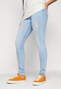Persona con jeans skinny rasgados de color azul claro y zapatillas naranja brillante con cordones, de pie contra un fondo blanco liso.