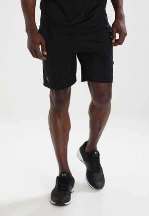 Uomo con pantaloncini sportivi neri, T-shirt nera e scarpe da ginnastica nero Under Armour, che cammina su uno sfondo bianco semplice.