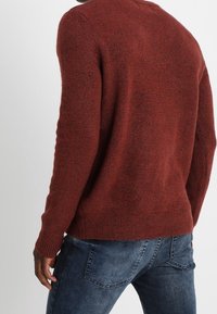 Maglione color ruggine con finitura a maglia testurizzata, collo rotondo e polsini a coste, abbinato a un paio di jeans in denim blu chiaro con tasche posteriori.