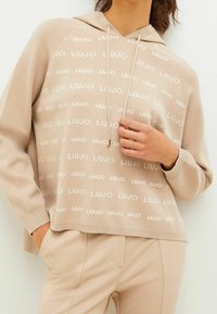 Beige hoodie met wit 'LIU JO' logo patroon, capuchon met trekkoord, geribbelde boorden en zoom. Gecombineerd met beige broek voor een coördinerende uitstraling.