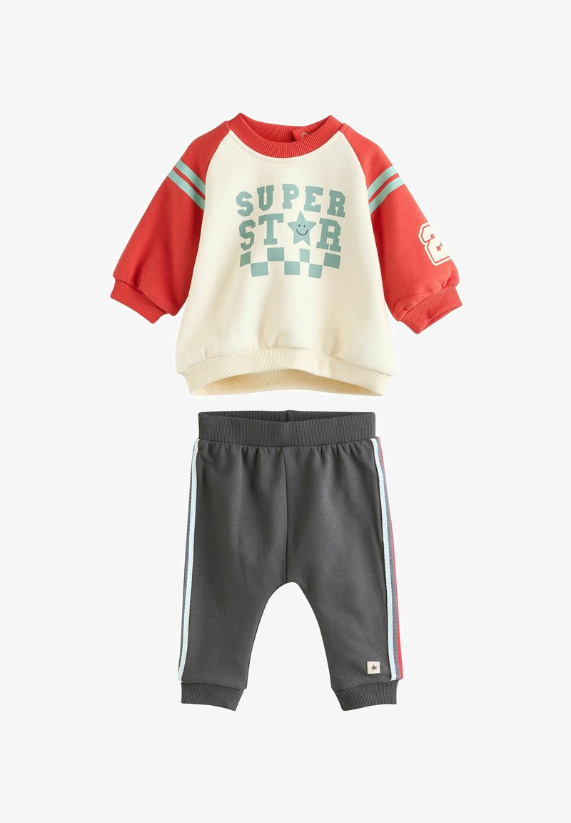 Krém és piros pulóver, teal csíkokkal és "SUPER STAR" grafikával; szürke melegítőnadrággal kombinálva, oldalcsíkokkal és gumírozott bokával.