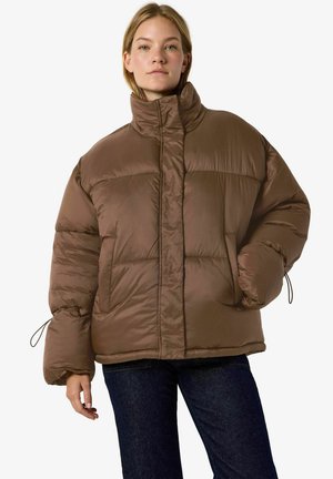 Une femme porte une doudoune marron oversize à col montant et un jean foncé, sur un fond blanc uni.