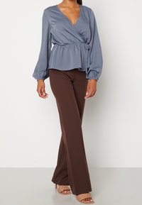 Kvinna i en blå långärmad omslagstopp, bruna höga vida byxor och beige vävda öppna klackar, stående mot en vit bakgrund.