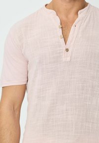 Chemise rose pâle à manches courtes en tissu texturé, avec une patte de boutonnage partielle munie de deux boutons en bois et un col mandarin.