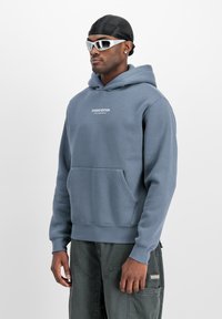 Hoodie blu in morbido tessuto, con tasca frontale e testo "STUDIO EDITION". Abbinato a pantaloni cargo grigi e occhiali da sole.