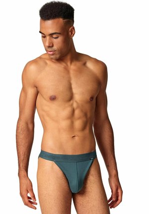 Male model with athletic build wearing dark green briefs, looking down and to his left, against white background.