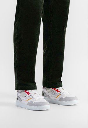 Zapatillas blancas y grises con detalles en rojo y dorado, con un upper texturizado y suela de goma plana, combinadas con pantalones de pana verde oscuro.