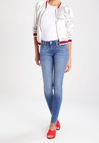 Chaqueta bomber plateada con rayas rojas y blancas en el cuello y los puños, combinada con jeans skinny azules y zapatillas rojas sin cordones.