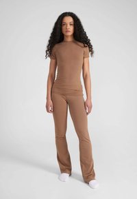 Bruin, aansluitend shirt met korte mouwen, gecombineerd met bijpassende flared broek, beide gemaakt van glad, rekbaar materiaal. Model staat op een effen achtergrond.