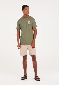 T-shirt in cotone verde oliva con una grafica colorata sul petto, abbinata a pantaloni corti beige e sandali neri. Design semplice e informale.