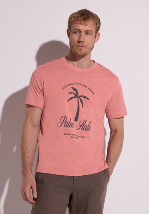 Uomo che indossa una T-shirt rosa corallo con grafica di palma e testo, abbinata a pantaloni marroni, in piedi davanti a uno sfondo chiaro e uniforme.