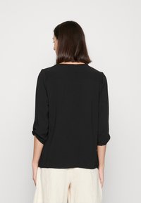 Schwarze, strukturierte Bluse mit Dreiviertelärmeln, die einen lässigen Schnitt und einen runden Halsausschnitt aufweist, kombiniert mit hellen, locker sitzenden Hosen.