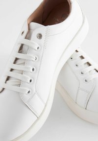 White leather sneakers with a smooth texture, round toe, six eyelets, and flat laces. Features subtle stitching and a contrasting sole.