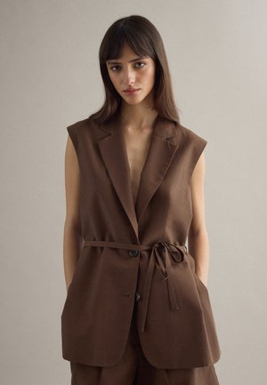 Femme portant un blazer marron sans manches avec ceinture nouée, debout devant un fond clair uni, mains dans les poches, regardant devant elle.
