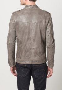 Giacca in pelle grigia con una texture liscia, dotata di epaulettes sulle spalle, dettagli di cucitura e una forma aderente. Indossata con jeans scuri.