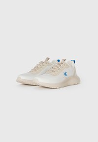Un par de zapatillas bajas en tonos beige y blanco con cordones beige, logotipo azul en los laterales y tiradores azules en los talones.
