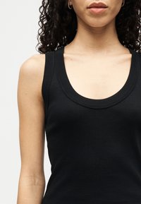 Femeie cu păr creț purtând un maiou fără mâneci negru cu dungi și cercei mici de tip cerc, prezentată de la umeri până la buze.