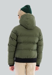 Persoon in een olijfgroene gewatteerde jas met capuchon, beige broek en een groene en beige gebreide muts, met de rug naar de camera tegen een lichtgrijze achtergrond.