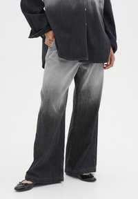 I pantaloni larghi in denim nero e grigio presentano una sfumatura gradiente, con una vestibilità comoda e cuciture visibili. Indossati con ballerine nere.