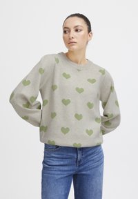 Pull en gris clair avec un motif de cœurs verts sur toute la surface, col rond et coupe décontractée. Associé à un jean bleu.