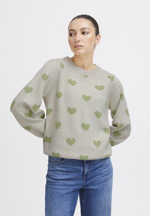 Pull en gris clair avec un motif de cœurs verts sur toute la surface, col rond et coupe décontractée. Associé à un jean bleu.