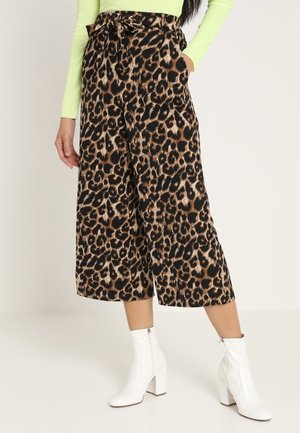 Vrouw draagt cropped broek met wijde pijpen en luipaardprint, neon groene top met lange mouwen en witte enkellaarzen met hak.