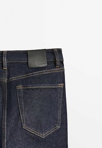 Ciemnoniebieskie dżinsy z czarną skórzaną metką marki nad tylną kieszenią, z złotymi szwami i klasycznym wzorem kieszeni.