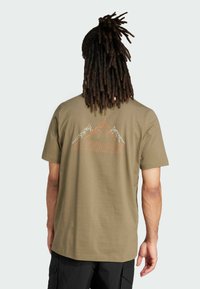 Oliven grøn bomulds t-shirt med et bjerggrafik i hvid og orange. Korte ærmer og en afslappet pasform. Logoet er vist på ryggen.