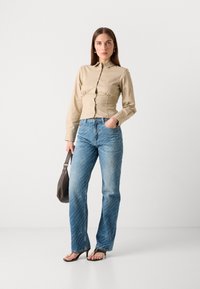 Camisa ajustada de color beige claro con cintura fruncida, combinada con jeans azules sueltos que presentan un sutil patrón blanco. La modelo sostiene un bolso marrón oscuro.