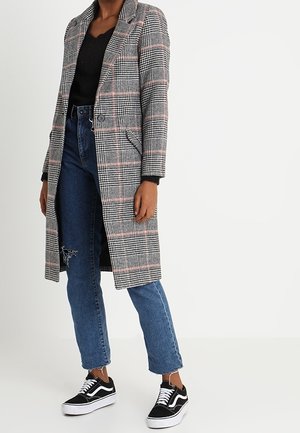 Cappotto grigio a quadri con accenti rosa, chiusura a pulsante singolo e collo a risvolto, indossato sopra un maglione nero e jeans blu, abbinato a sneakers nere.