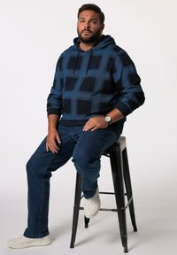 Felpa con cappuccio blu navy a fantasia plaid, indossata con jeans scuri e sneakers bianche, seduto su uno sgabello di metallo nero.