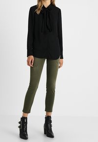 Svart långärmad blus med rosettkrage, ihopkopplad med åtsittande olivgröna jeans och kraftiga svarta klackstövlar med spänndetaljer.