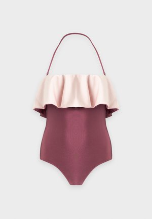 Une femme porte un maillot de bain une pièce couleur prune, orné d’un volant rose pâle sur la poitrine, devant un fond clair et uni.