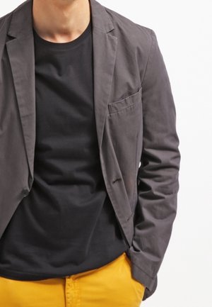 Mand iført en mørkegrå blazer over en sort t-shirt og sennepsgule bukser, stående med den ene hånd i lommen mod en hvid baggrund.