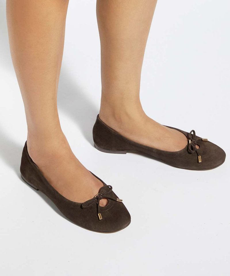 Pieds portant des ballerines en daim marron foncé avec de petites découpes à l'avant et des nœuds noués avec des embouts dorés sur fond blanc.