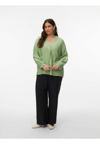 Blouse vert clair à manches longues avec col en V et détail plissé, portée avec un pantalon noir ample et des chaussures beiges sur un fond uni.