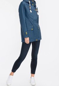 Sininen huppari, jossa on keltaisia nappeja ja valkoisia nyörejä, yhdistettynä tummiin slim fit -farkkuihin ja valkoisiin lenkkareihin seisovalla henkilöllä.