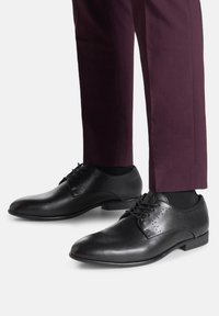 Czarne skórzane buty formalne z gładkim wykończeniem, delikatnymi perforacjami i zapinaniem na sznurowadła, w połączeniu z dopasowanymi spodniami w ciemnofioletowym kolorze.