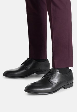 Juodos odinės suknelės bateliai su lygiu paviršiumi, subtiliomis perforacijomis ir raištelių užsegimu, derinami su tamsiai violetinėmis pritaikytomis kelnėmis.