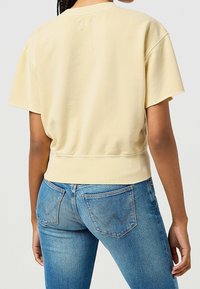 Frau, die ein hellgelbes kurzärmliges Sweatshirt und blaue Denim-Jeans trägt, von hinten vor einem schlichten hellen Hintergrund gezeigt.