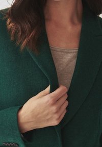 Cappotto verde in misto lana con collo a rever, mostrato leggermente aperto, che rivela una maglia beige chiaro sottostante. Texture liscia, dettaglio con singolo bottone.