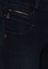Mörkblå denimjeans har en framficka med dragkedjestängning och en liten metallogotyp nära fickans söm.